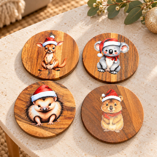 Four wooden Australian animal Christmas coasters displayed on a stone table beside festive eucalyptus and decorations.