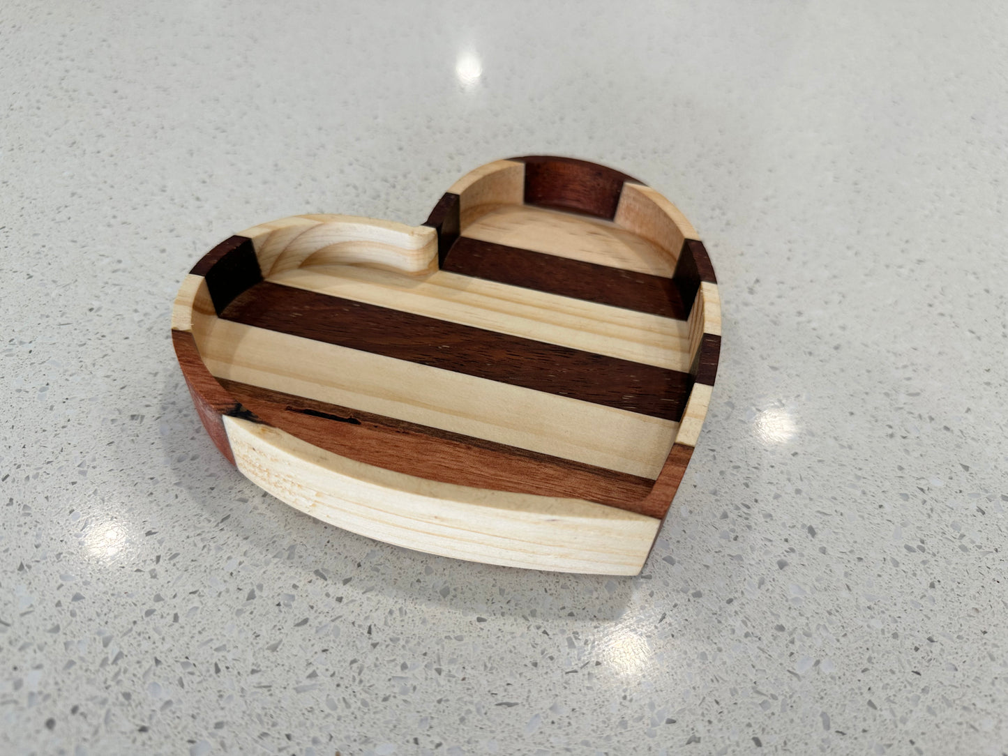 A heart-shaped reclaimed wood jewelry tray with a striped pattern, placed on a reflective surface.