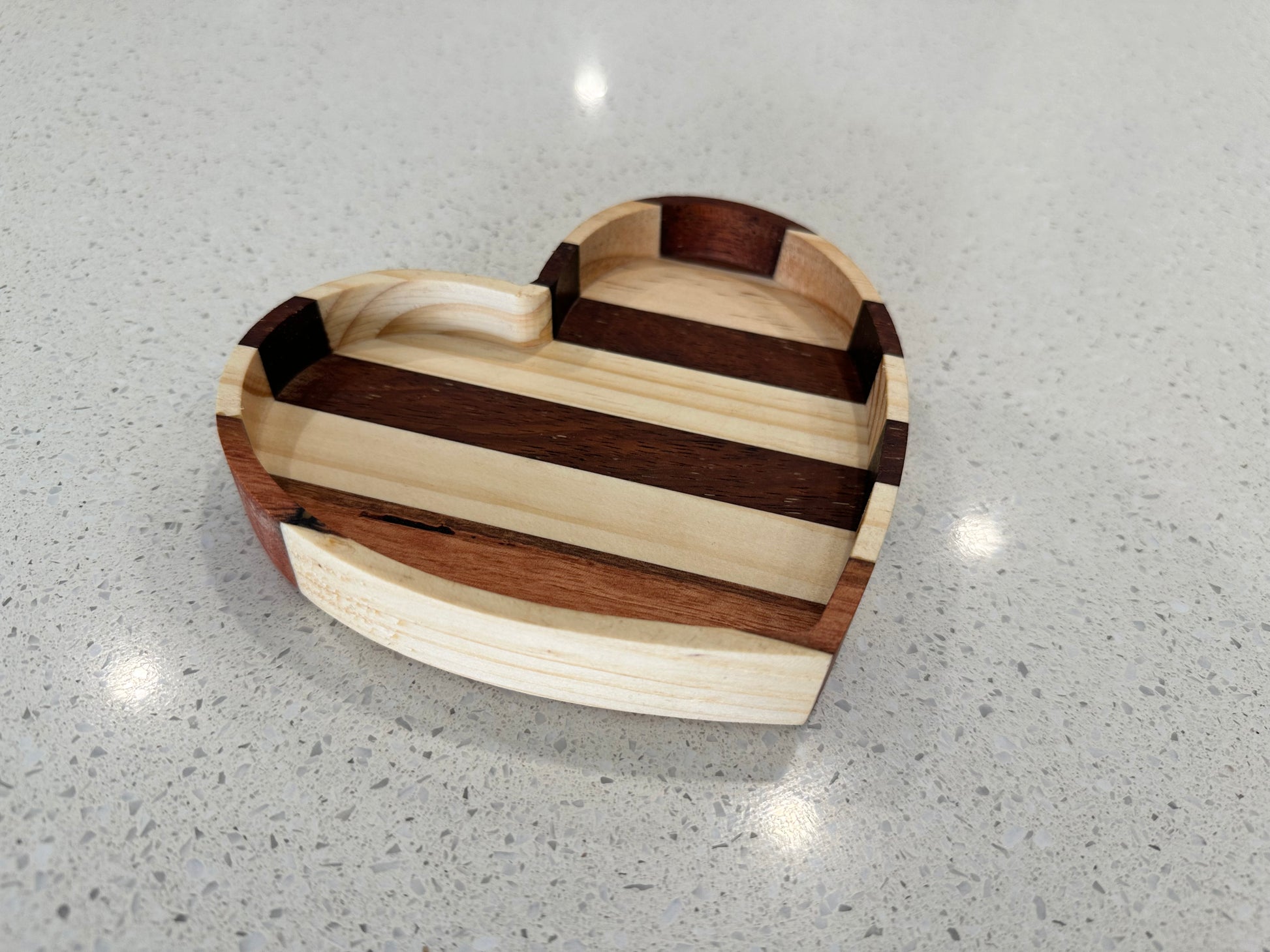 A heart-shaped reclaimed wood jewelry tray with a striped pattern, placed on a reflective surface.