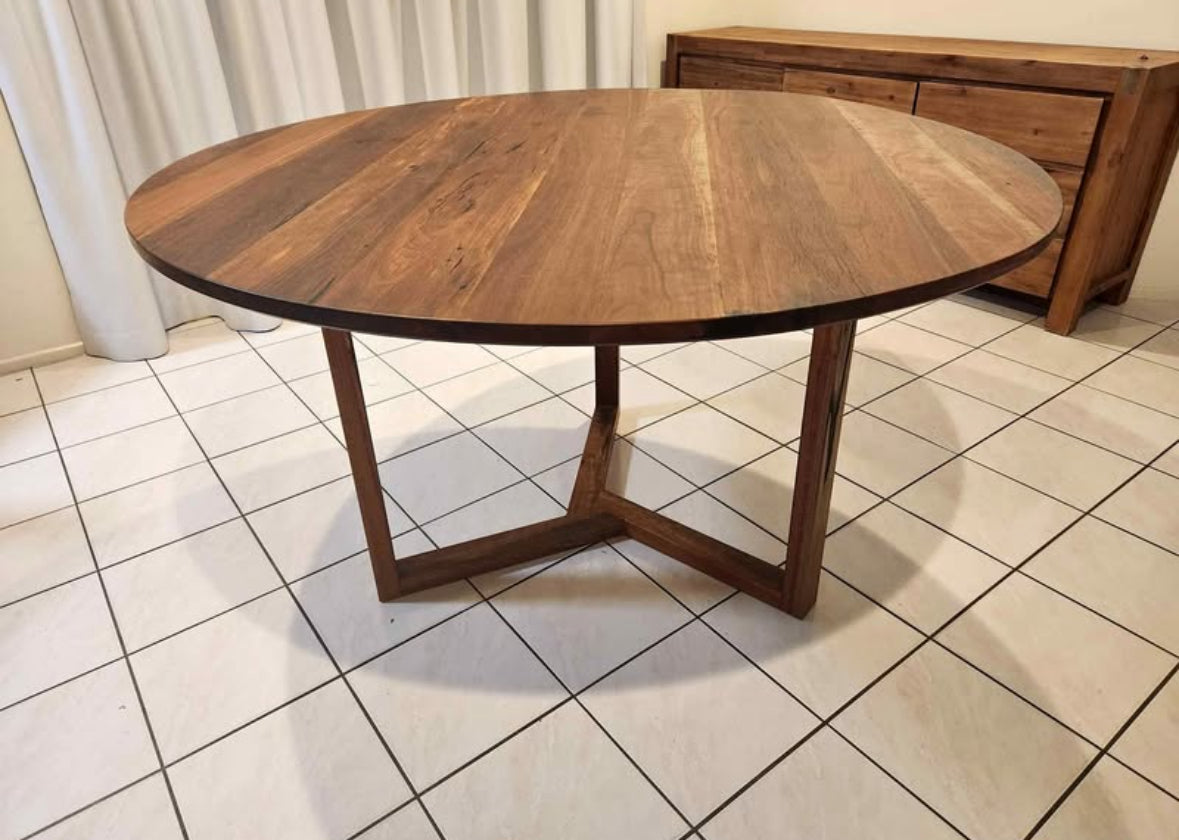 A round dining table made of Spotted Gum wood, featuring a tri-leg design, placed on a tiled floor.
